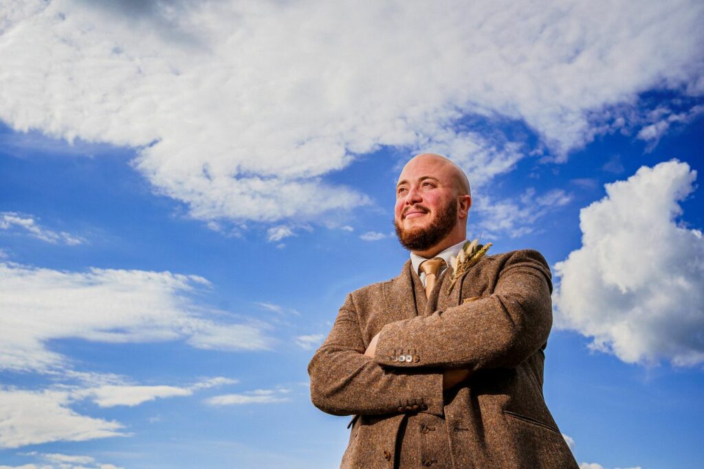 A bald man with a beard, wearing a brown tweed suit and tan tie, stands outdoors with his arms crossed, looking up and smiling. The sky behind him is bright blue with scattered white clouds. The sun casts a warm light on his face. Image by Documentary Wedding Photographer Lewis Bishop Photography.
