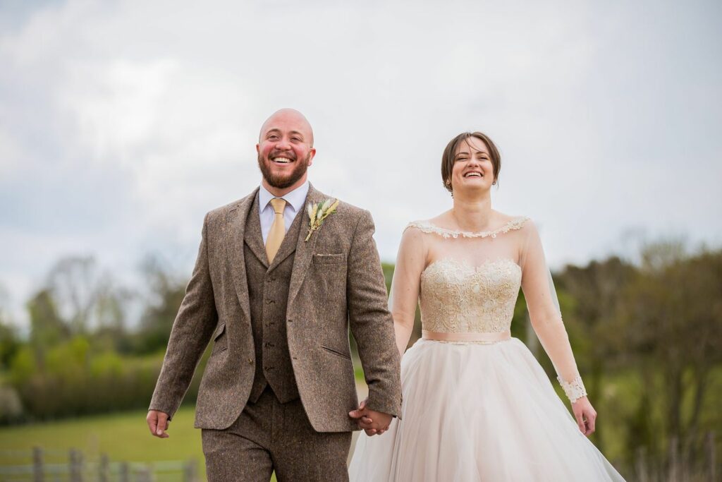 A couple dressed in wedding attire walks hand in hand outdoors. The man wears a brown tweed suit with a yellow boutonniere and the woman wears a white strapless wedding gown with a lace bodice. They both smile joyfully with a scenic green landscape in the background. Image by Documentary Wedding Photographer Lewis Bishop Photography.