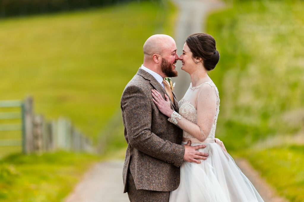 A bride and groom stand close together, touching noses and smiling broadly. The groom wears a brown suit and the bride is in a white wedding dress with lace details. Behind them, a path winds through a lush, green field under a clear sky. Image by Documentary Wedding Photographer Lewis Bishop Photography.