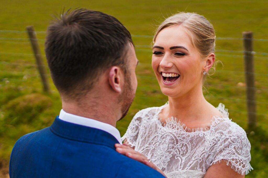 A bride in a white lace dress and a groom in a blue suit share a joyful moment outdoors in front of a green field and wire fence. The bride is smiling widely, looking at the groom, who has his back to the camera. Image by Documentary Wedding Photographer Lewis Bishop Photography.