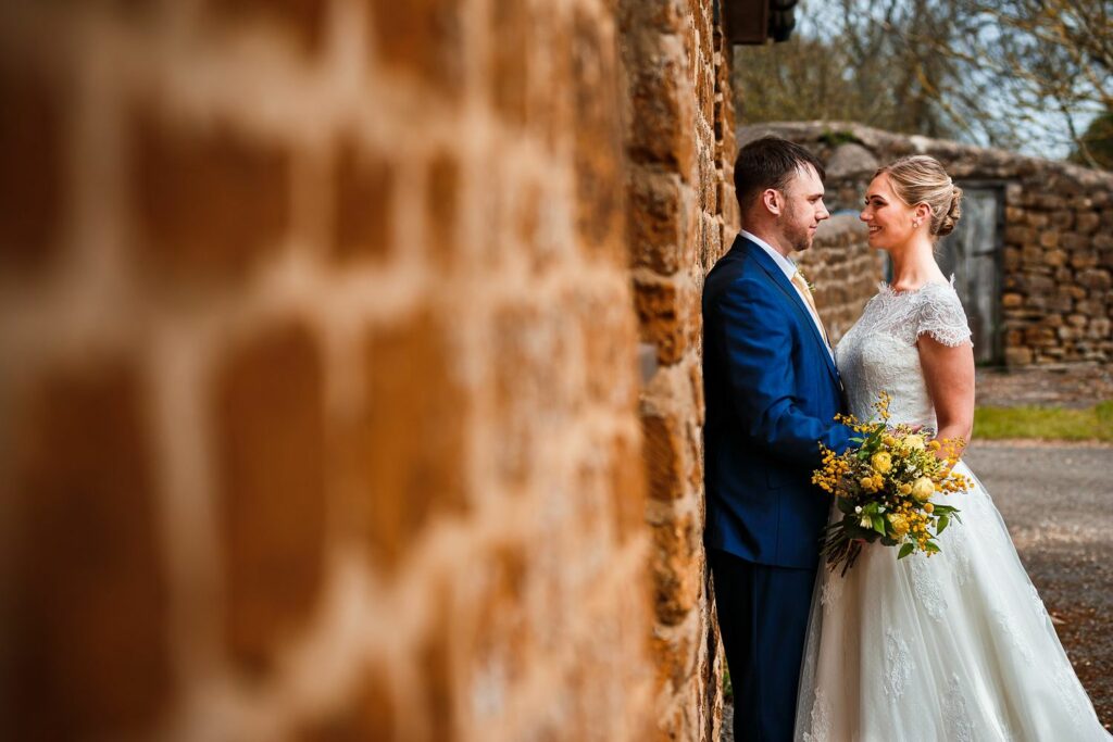A bride in a white lace wedding dress and a groom in a navy suit stand face-to-face near a rustic stone wall. The bride holds a bouquet of yellow flowers, and they gaze at each other lovingly. The background includes more stone structures and trees. Image by Documentary Wedding Photographer Lewis Bishop Photography.