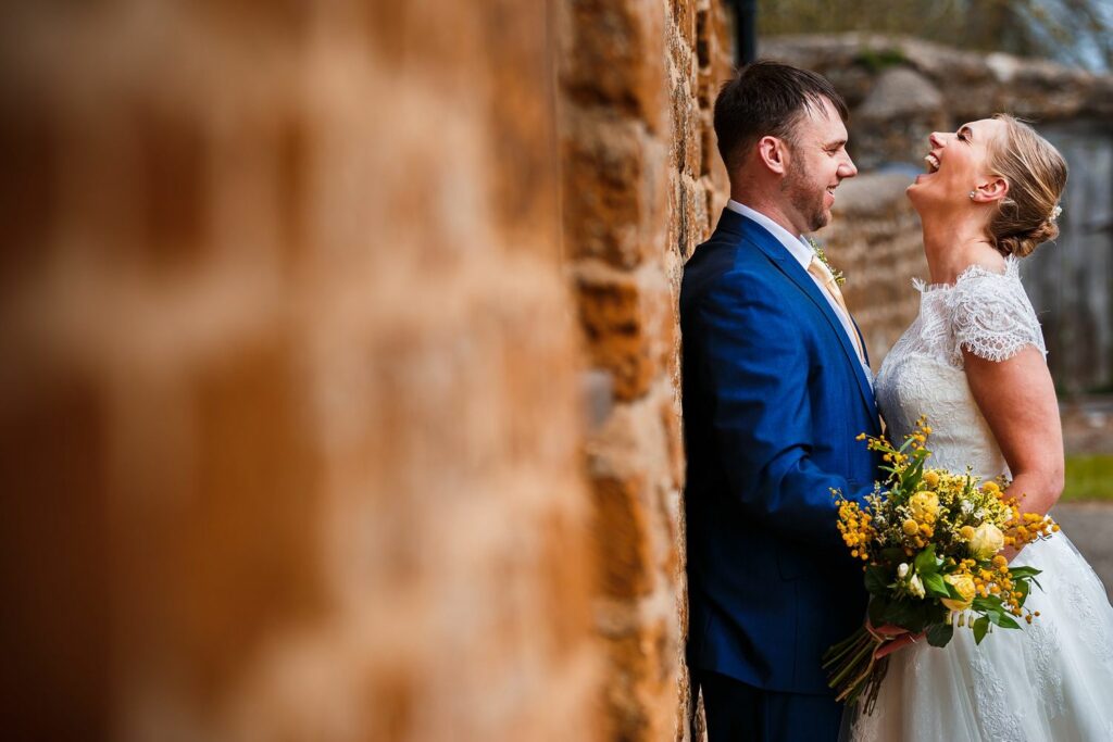 A bride and groom share a joyful moment, standing close together by a rustic brick wall. The bride is in a white lace dress holding a yellow bouquet, and the groom is in a blue suit. Both are smiling and looking at each other, exuding happiness. Image by Documentary Wedding Photographer Lewis Bishop Photography.