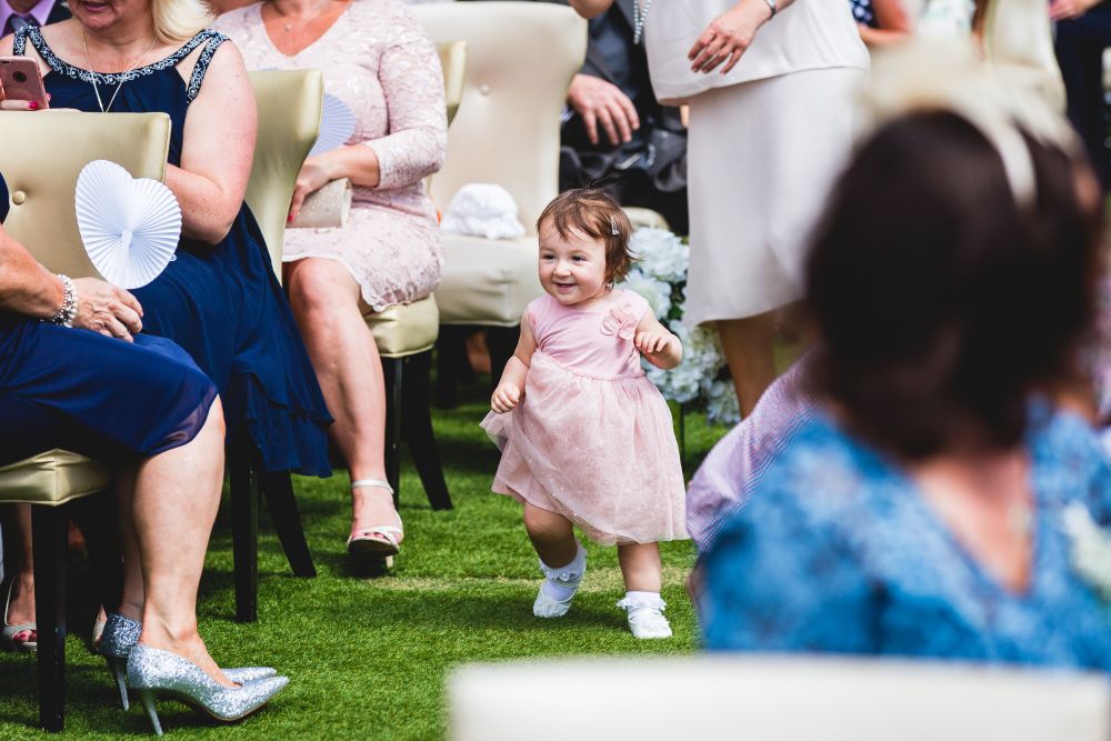 A young child in a pink dress and white shoes joyfully walks down the aisle outside, surrounded by seated adults dressed in formal attire. The event appears to be a wedding or a formal gathering, with people smiling and watching the child. Image by Documentary Wedding Photographer Lewis Bishop Photography.
