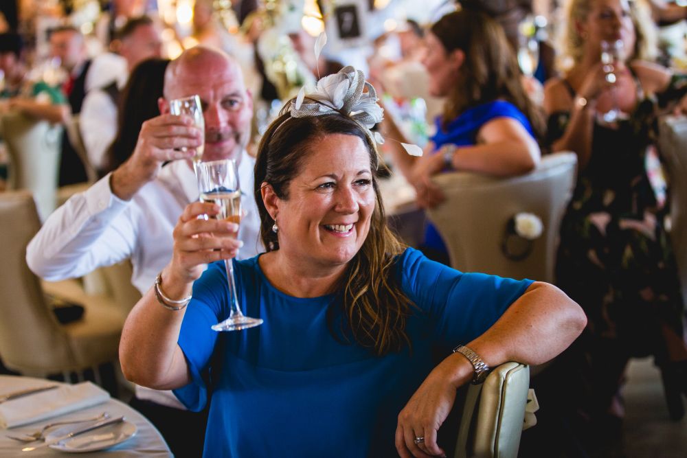 A woman in a blue dress raises a glass and smiles amid a crowded festive gathering. Beside her, a man also holds up a glass in a toast. Other guests in the background appear to be engaged in the celebration. The atmosphere is joyful and lively. Image by Documentary Wedding Photographer Lewis Bishop Photography.