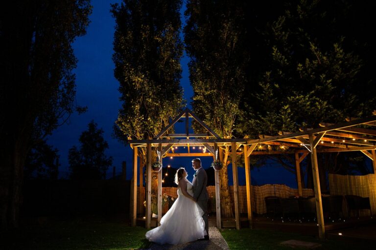 A newlywed couple stands under a wooden pergola adorned with string lights. The bride, in a voluminous white dress, gazes at the groom. Tall trees and a dark, starless night sky form the backdrop, creating a romantic and serene atmosphere. Image by Documentary Wedding Photographer Lewis Bishop Photography.