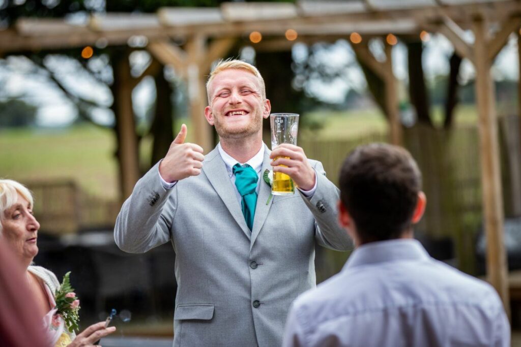 A smiling man in a light gray suit and teal tie gives a thumbs-up while holding a glass of beer. He appears to be at an outdoor event or celebration with blurred people and a wooden pergola in the background. Image by Documentary Wedding Photographer Lewis Bishop Photography.