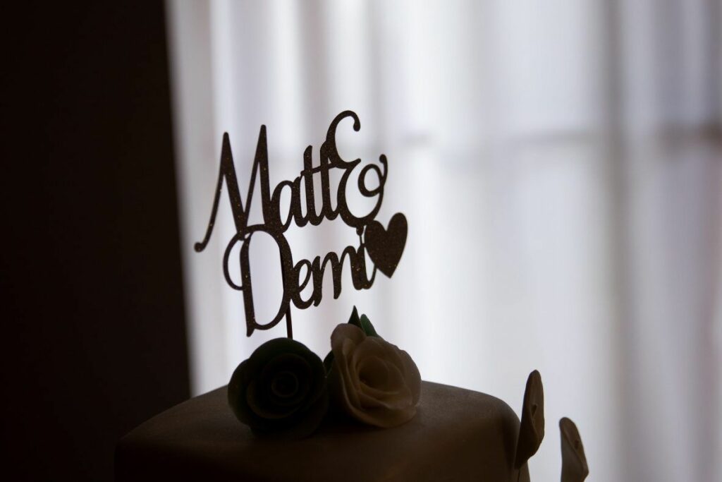 A wedding cake topper featuring the names "Matt & Demi" in cursive script with a heart shape sits atop a wedding cake. The cake is decorated with green and white fondant roses against a softly lit background. Image by Documentary Wedding Photographer Lewis Bishop Photography.
