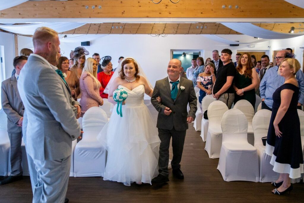 A bride in a white dress walks down the aisle with an older man in a grey suit. Guests stand on both sides, watching her approach a bearded man in a light grey suit at the altar. The room is decorated with white chairs and overhead drapery. Image by Documentary Wedding Photographer Lewis Bishop Photography.