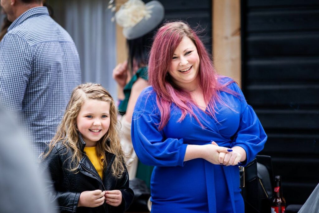 A smiling woman with pink hair in a blue dress stands beside a young girl with wavy blonde hair wearing a black leather jacket and yellow shirt. They appear to be enjoying an event with other people in the background. Image by Documentary Wedding Photographer Lewis Bishop Photography.