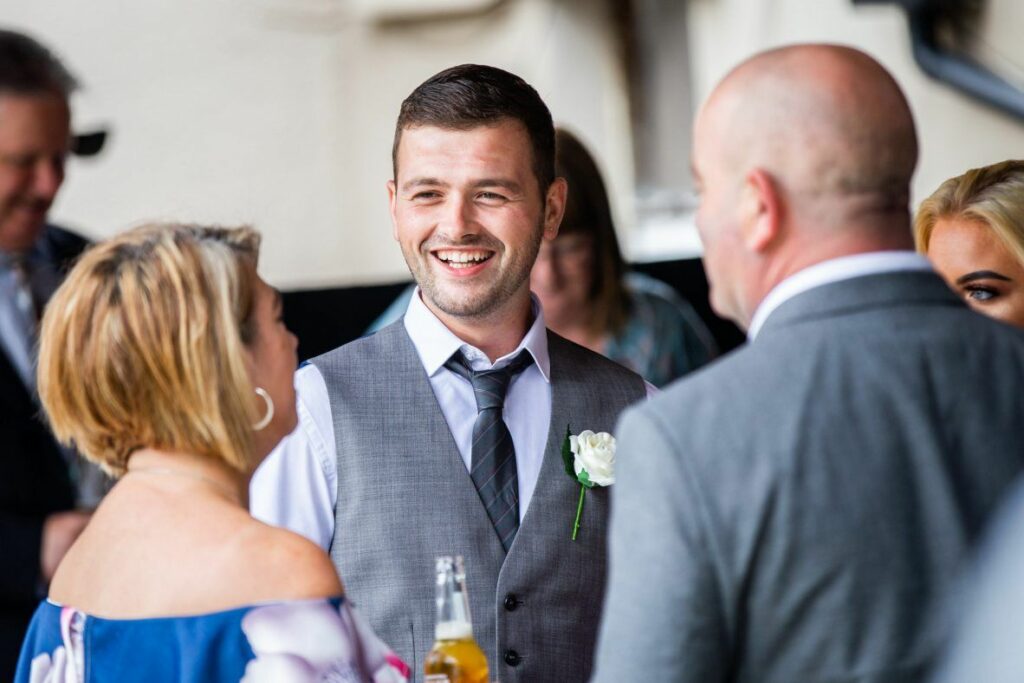 A man in a gray vest and plaid tie, holding a beer, smiles while talking to guests at an event. He has a white boutonnière on his vest. A woman in an off-the-shoulder floral dress and another man in a gray suit are also engaged in the conversation. Image by Documentary Wedding Photographer Lewis Bishop Photography.