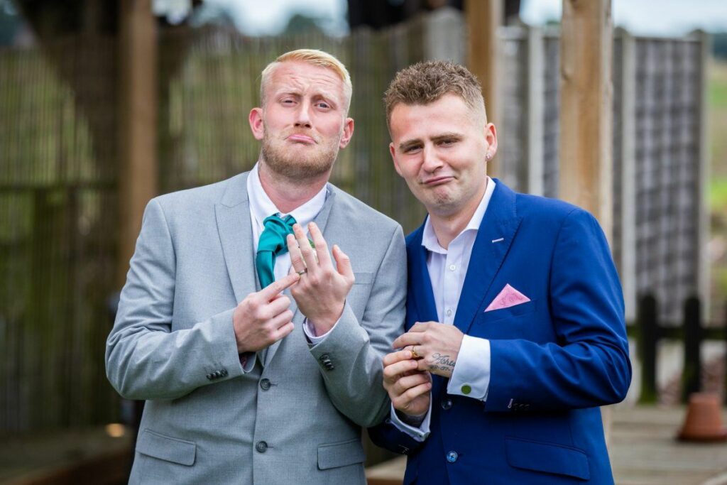 Two men in suits are standing outdoors, each holding up their hands to display rings on their ring fingers. The man on the left is wearing a light grey suit with a teal tie, and the man on the right is wearing a dark blue suit with a pink handkerchief. Both are looking at the camera. Image by Documentary Wedding Photographer Lewis Bishop Photography.