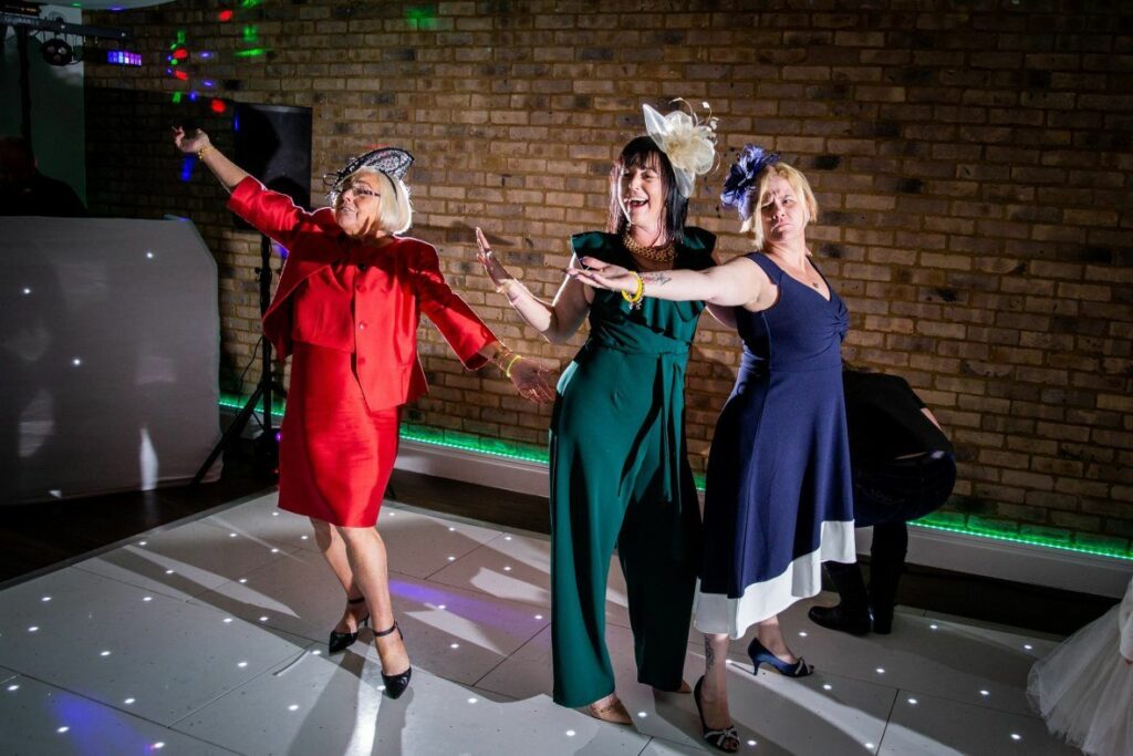 Three women in vibrant outfits, including a red dress, green jumpsuit, and blue dress, joyfully dance together on a white-lit dance floor. They wear stylish fascinators and appear to be celebrating, with a colorful brick wall and a DJ booth in the background. Image by Documentary Wedding Photographer Lewis Bishop Photography.