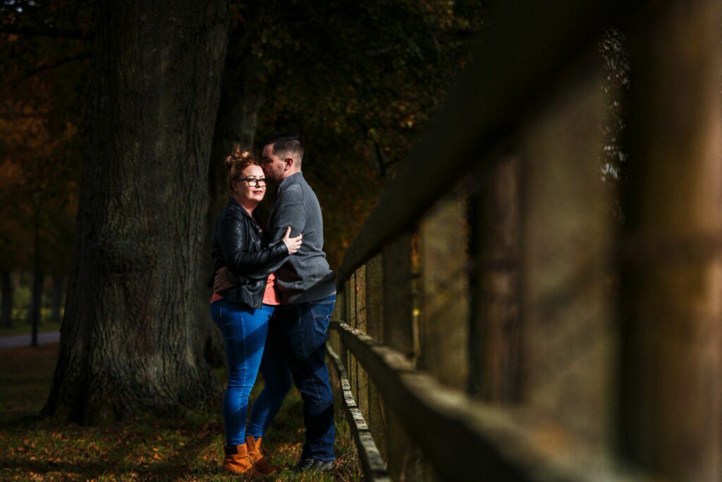 A couple stands close together, smiling and embracing near a wooden fence in a wooded area. The woman wears a black jacket and jeans, and the man is in a gray jacket and jeans. The background features tall trees and dappled sunlight filtering through the leaves. Image by Documentary Wedding Photographer Lewis Bishop Photography.