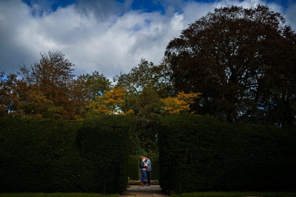 A couple stands closely together in front of a large hedge with a passageway, surrounded by trees with autumn foliage under a partly cloudy sky. They appear to be enjoying a private moment in the scenic garden. Image by Documentary Wedding Photographer Lewis Bishop Photography.