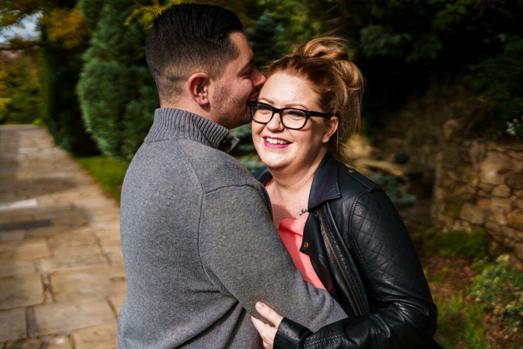 A smiling woman with glasses and a black leather jacket embraces a man in a gray sweater who is whispering in her ear. They are outdoors on a sunny day with greenery and a stone pathway in the background. Image by Documentary Wedding Photographer Lewis Bishop Photography.
