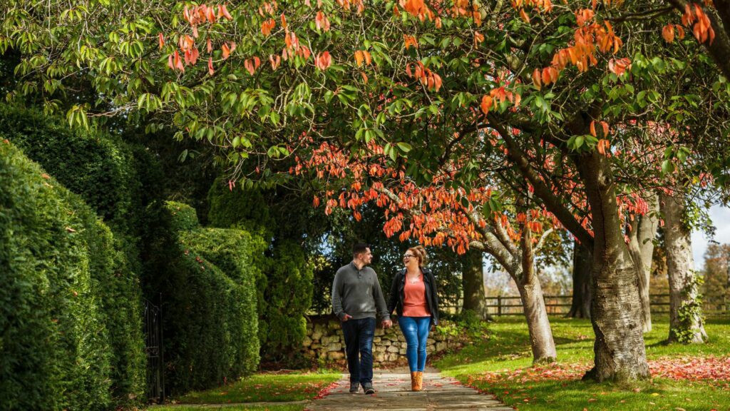 A couple walks hand in hand on a stone pathway, surrounded by lush greenery and trees with autumn foliage. They are under a canopy of leaves in shades of orange and green. The scene is serene with a stone wall in the background. Image by Documentary Wedding Photographer Lewis Bishop Photography.
