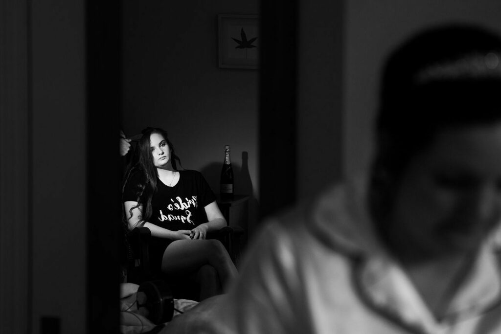 A black-and-white photo captures a woman sitting and looking thoughtfully to the side, wearing a shirt that reads "Bride Squad." In the blurred foreground, another person is seen, possibly preparing for an event. A bottle is visible in the background on a table. Image by Documentary Wedding Photographer Lewis Bishop Photography.