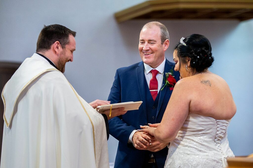 A bride and groom stand facing each other, smiling, as they exchange rings in a wedding ceremony. The officiant, wearing a white robe, is beside them holding a book and also smiling. The bride wears a strapless white gown, and the groom is in a blue suit with a red tie. Image by Documentary Wedding Photographer Lewis Bishop Photography.