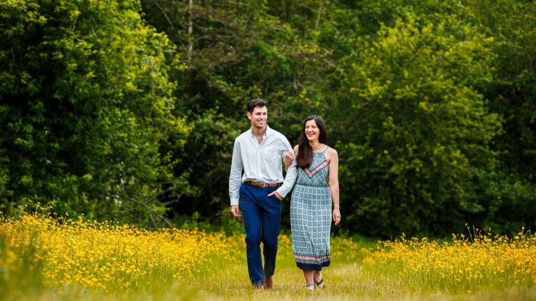 A man and a woman walk arm in arm through a vibrant field of yellow flowers. Both are smiling and appear happy in the lush, green outdoor setting. The man is wearing a white shirt and blue pants, while the woman is in a patterned dress. Trees are visible in the background. Image by Documentary Wedding Photographer Lewis Bishop Photography.