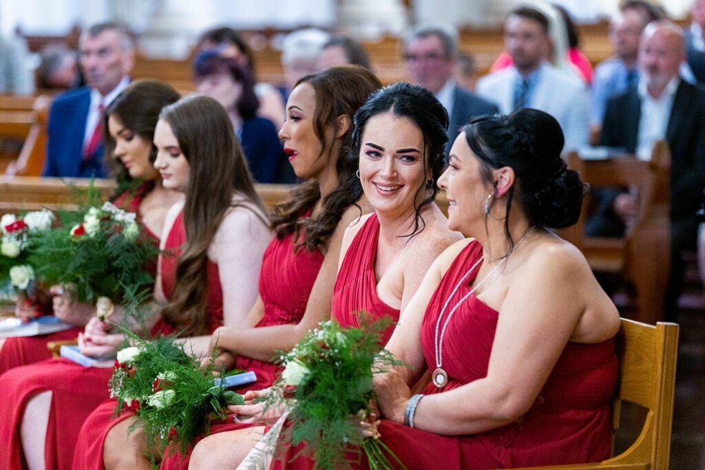 Four bridesmaids in red dresses sit in a church pew, holding green floral bouquets. Some smile and talk amongst themselves. The background shows seated guests, out of focus, in the church. Image by Documentary Wedding Photographer Lewis Bishop Photography.