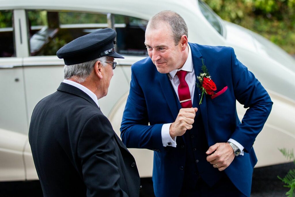 A man in a blue suit with a red tie and boutonnière leans in while speaking to an older man in a black uniform and cap. They are standing outside near a vintage white car. Both men appear to be engaged in friendly conversation. Image by Documentary Wedding Photographer Lewis Bishop Photography.