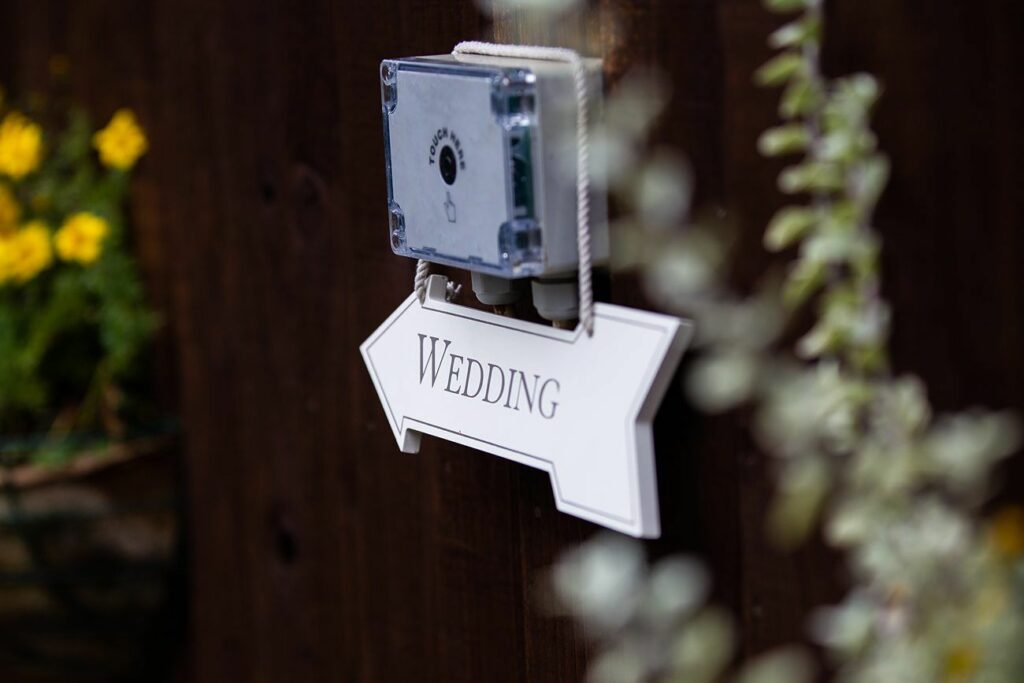 A wedding sign in the shape of an arrow is mounted on a dark wooden wall with a small, transparent electronic device above it. Yellow flowers and some greenery are blurred in the background. The sign points to the right. Image by Documentary Wedding Photographer Lewis Bishop Photography.