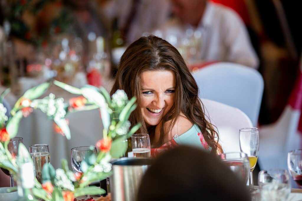 A woman with long brown hair is laughing while seated at a table during a formal event. She is surrounded by glasses and decorations, including green and red floral arrangements. People in the background are blurred but appear to be enjoying the occasion. Image by Documentary Wedding Photographer Lewis Bishop Photography.