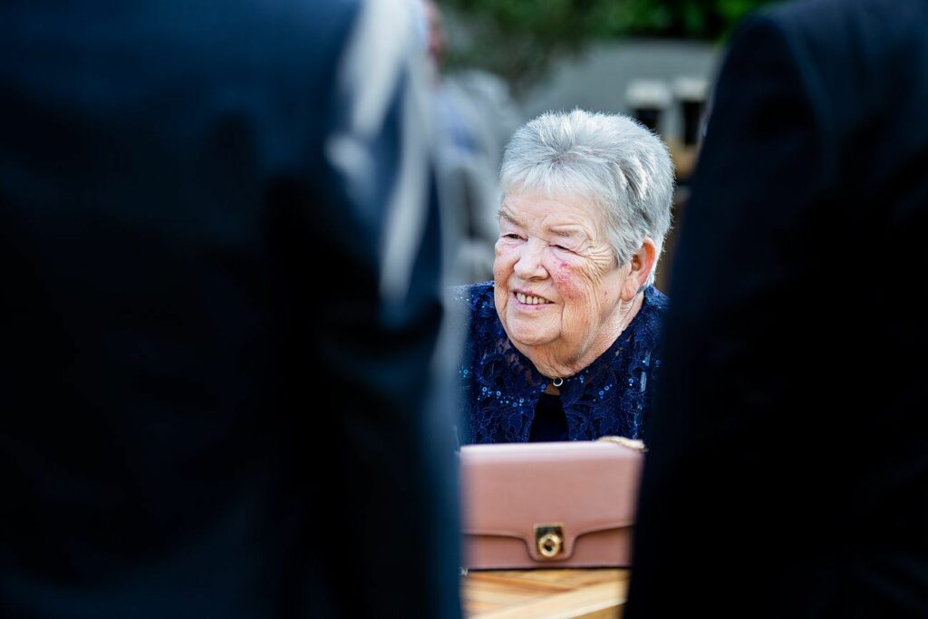 An elderly woman with short gray hair and a smile on her face is seated at a table outdoors. She is wearing a dark blue outfit and holding a beige handbag. The photo is focused on her, with two people in darker outfits partially visible in the foreground. Image by Documentary Wedding Photographer Lewis Bishop Photography.