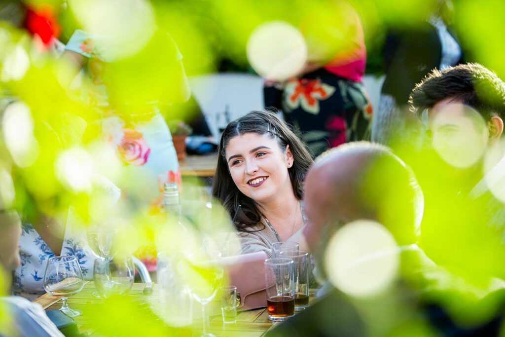A young woman with dark hair smiles while sitting at an outdoor gathering. She is surrounded by other people and tables with drinks. The image is creatively framed with green leaves in the foreground, adding a vibrant, natural border. Image by Documentary Wedding Photographer Lewis Bishop Photography.