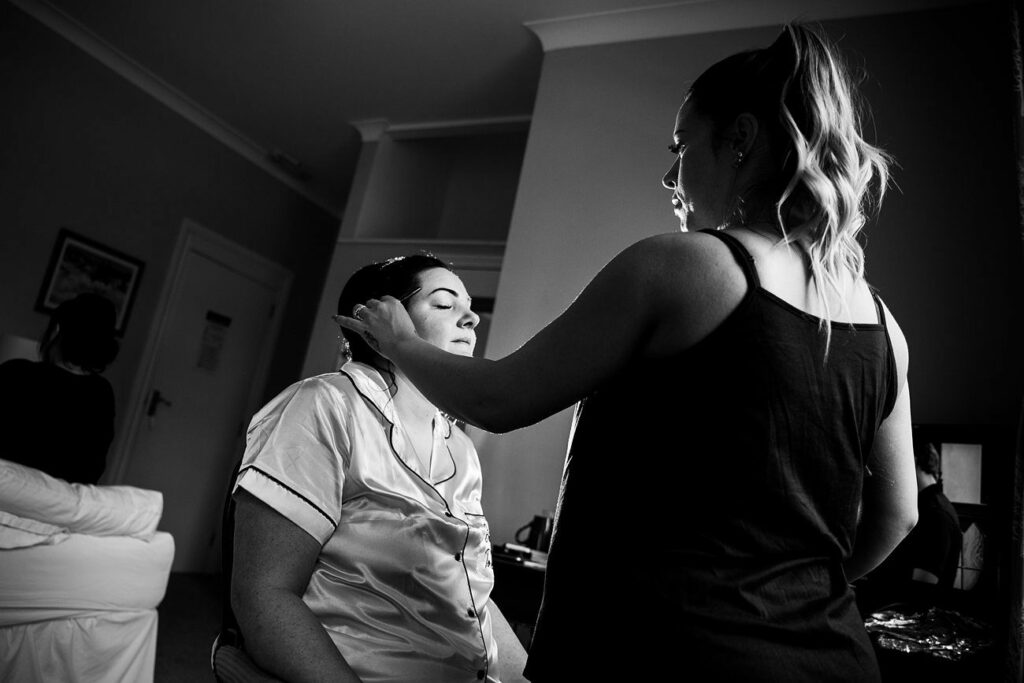 A woman with closed eyes sits in a chair, while another woman stands in front of her, applying makeup. The room has soft lighting and a relaxed atmosphere. Another person is distantly visible in the background. Black and white photo. Image by Documentary Wedding Photographer Lewis Bishop Photography.