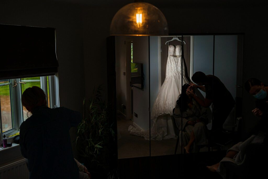 A dimly lit room shows a bride getting her makeup done. She sits in front of a mirror where her reflection, along with the reflection of her wedding dress on a hanger, is visible. A makeup artist is applying makeup, and another person stands nearby, observing. Image by Documentary Wedding Photographer Lewis Bishop Photography.