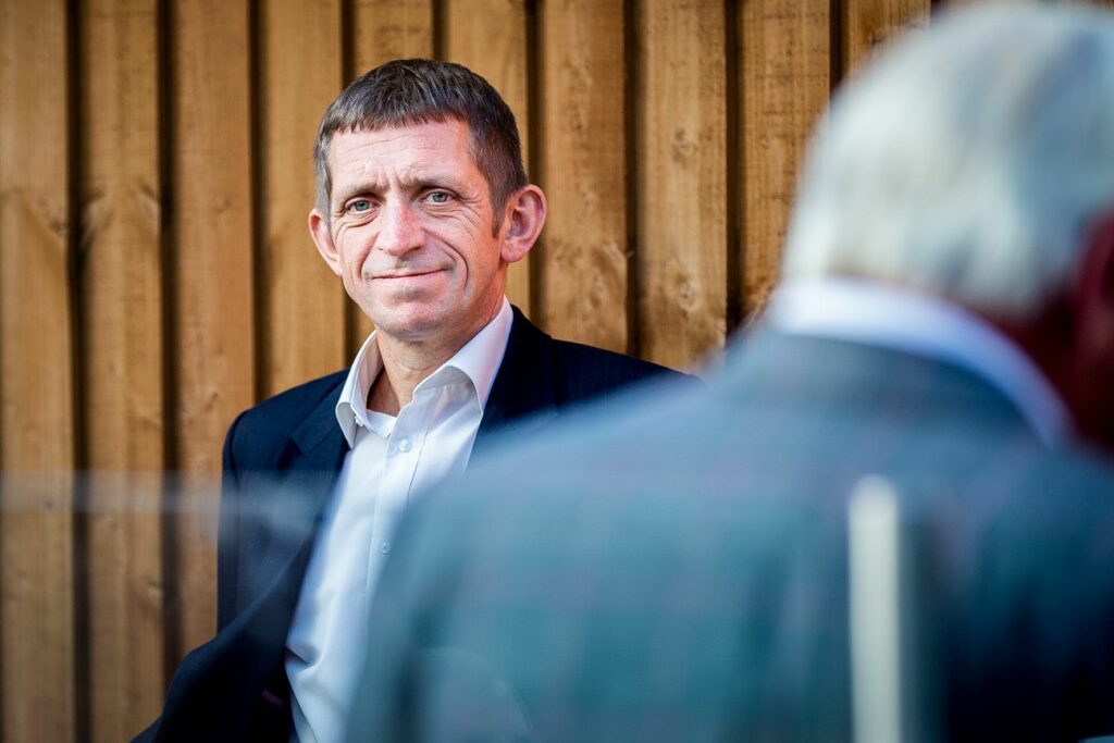 A man in a suit and white shirt is standing against a wooden wall, looking at the camera. The background features another man facing away, blurred in the foreground, with gray hair and also dressed in a suit. Image by Documentary Wedding Photographer Lewis Bishop Photography.