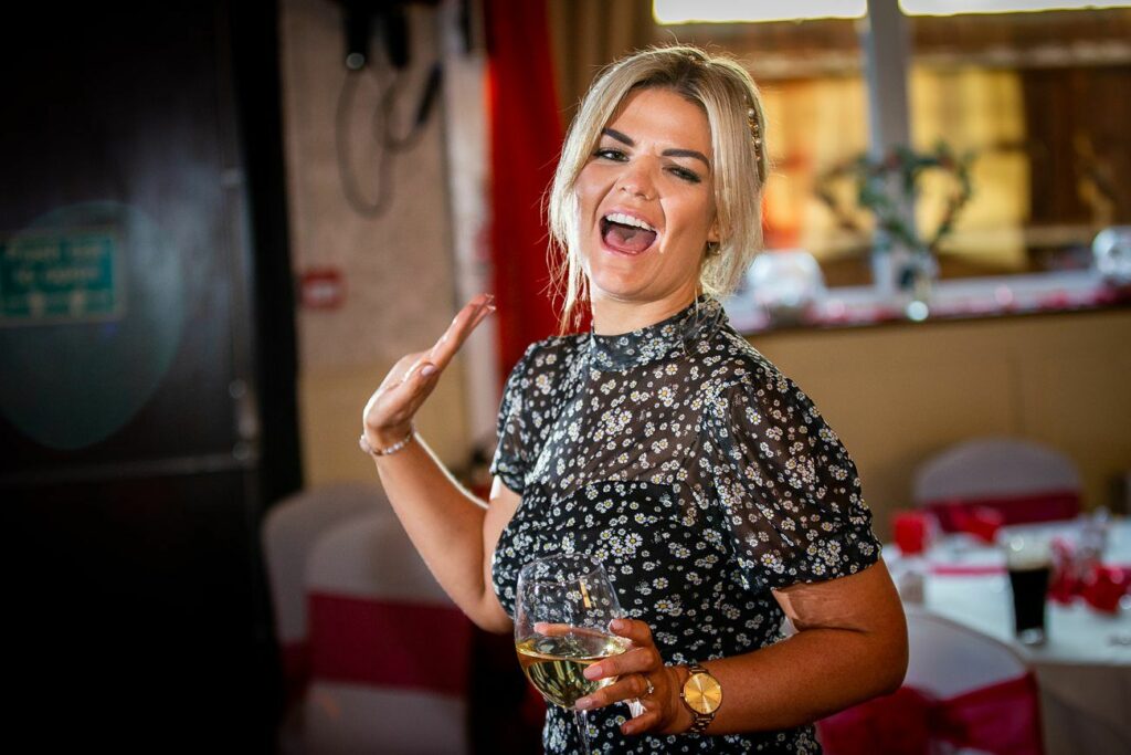 A woman with blonde hair smiling warmly waves with one hand while holding a glass of white wine in the other. She is dressed in a black floral top and standing in a brightly lit room with red and white decorations. Image by Documentary Wedding Photographer Lewis Bishop Photography.