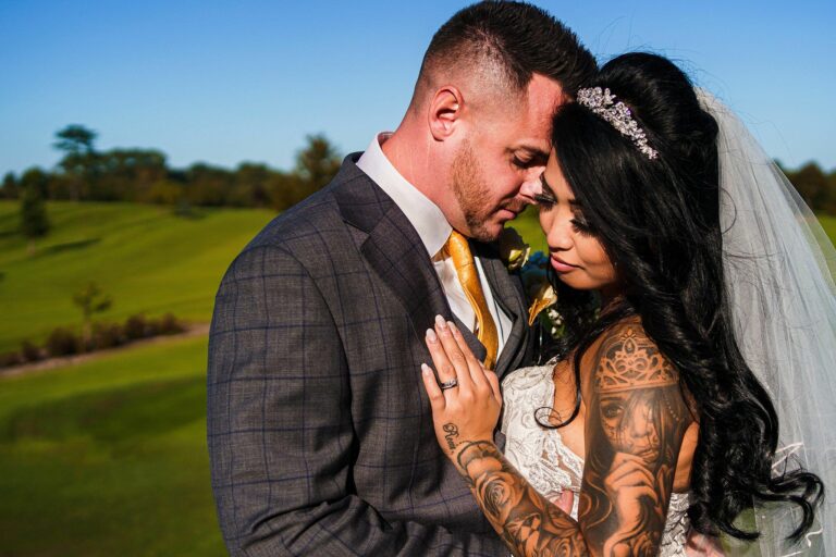 A bride and groom share an intimate moment on their wedding day. They are standing close together with their foreheads touching. The groom is wearing a checked suit with a yellow tie, and the bride is dressed in a white gown with visible tattoos and a headpiece. The background is an open, grassy field. Image by Documentary Wedding Photographer Lewis Bishop Photography.