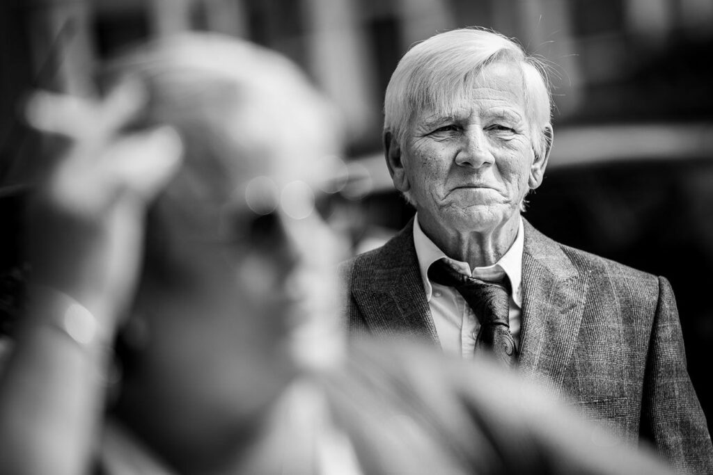 Black-and-white photo of an elderly man with white hair, wearing a suit and tie, standing outdoors. He looks ahead with a serious expression. In the blurred foreground, a person with short hair, wearing glasses, is seen touching their head. Image by Documentary Wedding Photographer Lewis Bishop Photography.