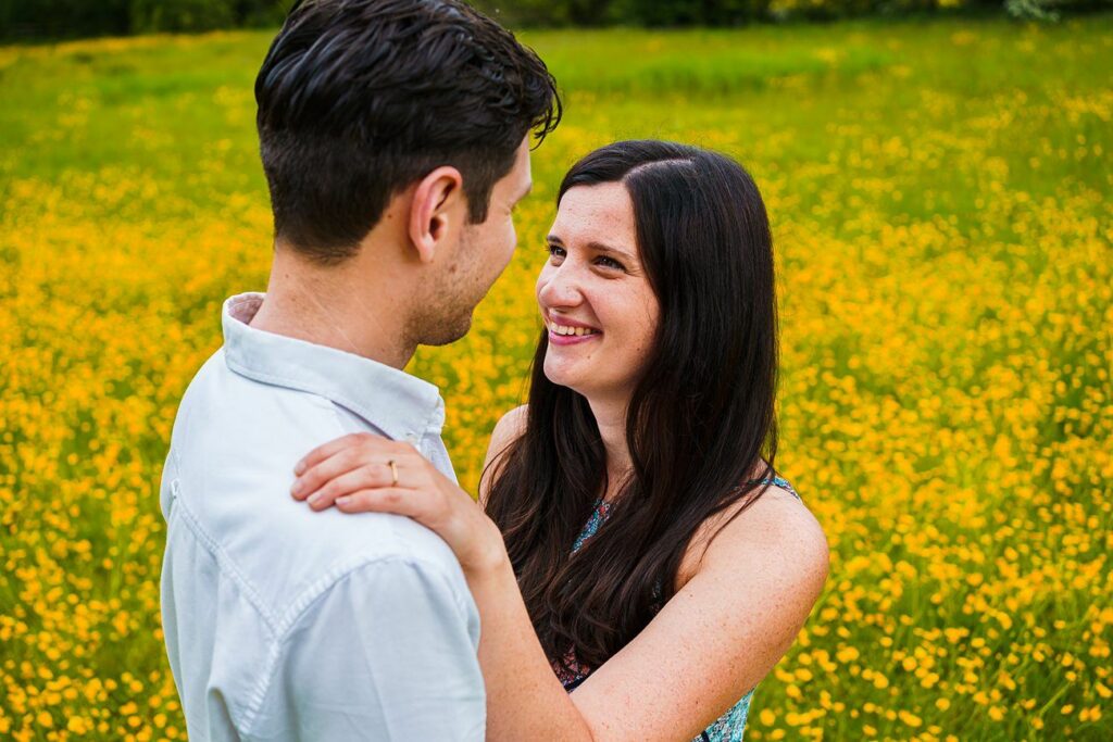 A couple stands in a field of yellow flowers. The woman has long dark hair and is smiling while placing her hand on the shoulder of the man, who has short dark hair. They are looking into each other's eyes, creating a joyful and intimate moment. Image by Documentary Wedding Photographer Lewis Bishop Photography.