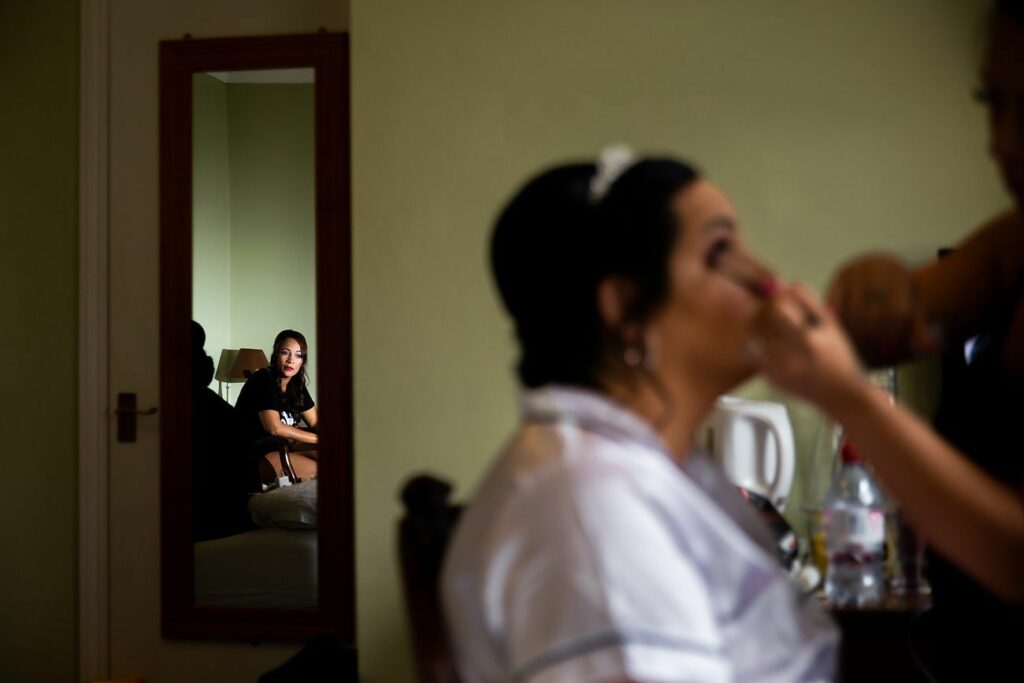 A woman is getting her makeup done by another person in the foreground. In the background, her reflection is visible in a full-length mirror, showing another woman sitting on a bed and smiling. The room has neutral-colored walls and a wooden door. Image by Documentary Wedding Photographer Lewis Bishop Photography.