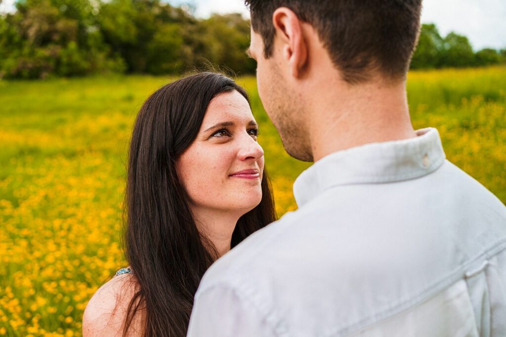 A woman with long dark hair smiles warmly at a man with short dark hair. They are standing close together in a field of yellow flowers with green trees in the background, creating a serene, nature-filled setting. Image by Documentary Wedding Photographer Lewis Bishop Photography.