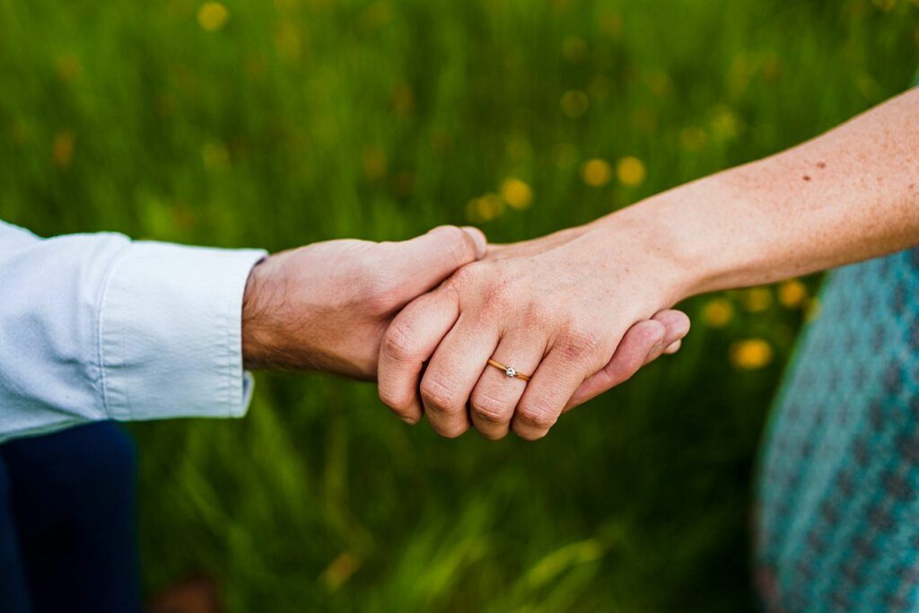 Close-up of two people holding hands, with one wearing an engagement ring. The background is a blurred field of green grass and yellow flowers. One person is dressed in a white long-sleeve shirt, while the other is wearing a turquoise patterned outfit. Image by Documentary Wedding Photographer Lewis Bishop Photography.