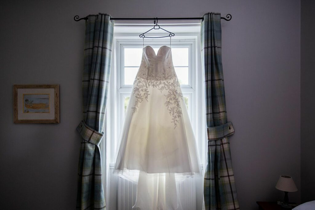 A white wedding dress with intricate lace details hangs on a hanger in front of a window framed by blue and gray plaid curtains. A small lampshade and a framed painting are visible on either side of the window. Image by Documentary Wedding Photographer Lewis Bishop Photography.