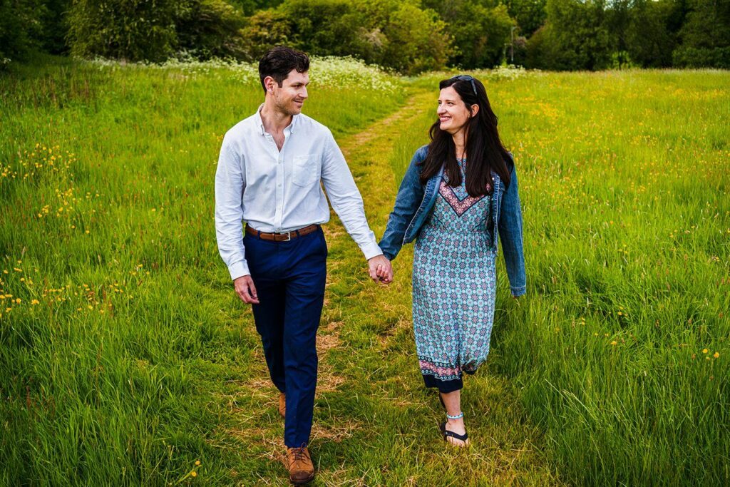 A man and a woman are walking hand in hand through a lush green field with tall grass and wildflowers. The man is wearing a white shirt and blue pants, and the woman is wearing a blue patterned dress and a denim jacket. Both are smiling and looking at each other. Image by Documentary Wedding Photographer Lewis Bishop Photography.