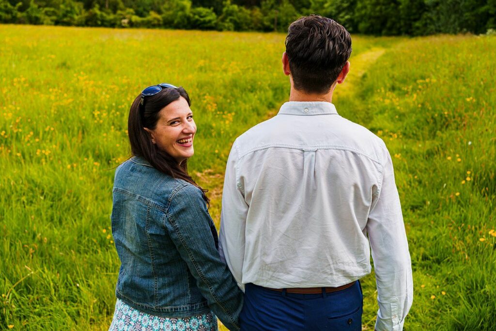 A couple stands in a lush green field with yellow flowers, holding hands. The woman, wearing a denim jacket and a floral dress, smiles and looks back over her shoulder at the camera. The man, in a white shirt and blue pants, faces away from the camera. Image by Documentary Wedding Photographer Lewis Bishop Photography.