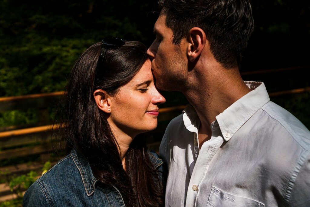 A man gently kisses a woman's forehead in an outdoor setting. The woman has her eyes closed and smiles contentedly. They both have dark hair; she wears a denim jacket, and he is in a white button-up shirt. Green foliage and a wooden fence are in the background. Image by Documentary Wedding Photographer Lewis Bishop Photography.