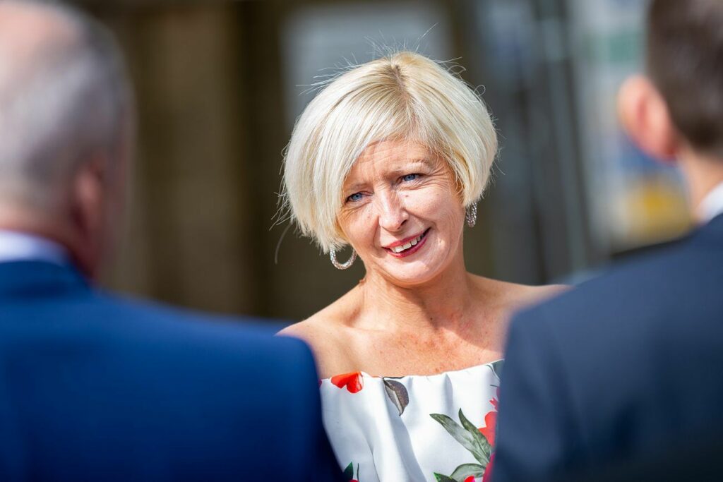 A woman with short blonde hair, wearing a white off-the-shoulder dress with a floral pattern, smiles and engages in conversation with two people. The background is blurred, focusing on her expressive smile and friendly demeanor. Image by Documentary Wedding Photographer Lewis Bishop Photography.