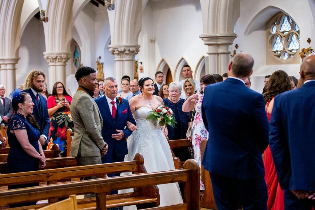 A bride in a white gown walks down the aisle of a church, accompanied by a man in a gray suit. Guests, some seated and some standing, look on with smiles. The church has ornate arches and stained glass windows. The atmosphere is joyful. Image by Documentary Wedding Photographer Lewis Bishop Photography.