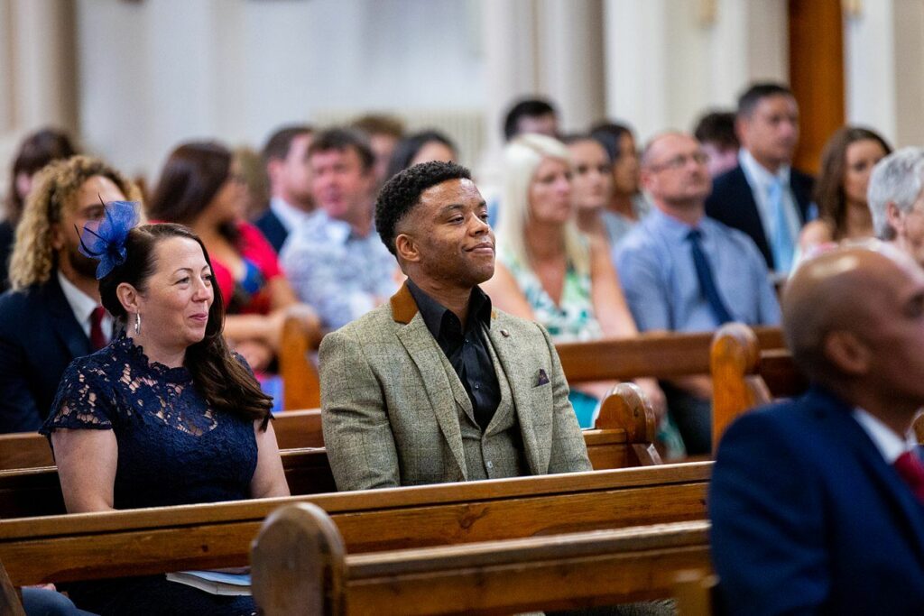 A group of people sit on wooden pews inside a church, dressed formally. A man in a gray suit and a woman in a navy blue dress with a fascinator are in the foreground, smiling and looking ahead. The background features other attendees, some looking forward and others conversing. Image by Documentary Wedding Photographer Lewis Bishop Photography.