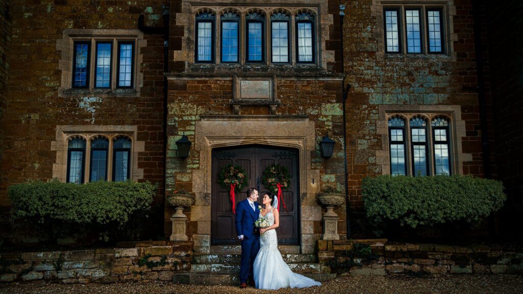 A bride in a white gown and a groom in a blue suit stand closely together in front of a grand, rustic building adorned with large windows and greenery. The entrance behind them is framed by two wreaths, adding a festive touch to the scene. Image by Documentary Wedding Photographer Lewis Bishop Photography.