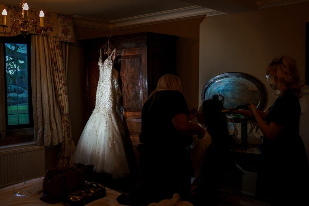 A dimly lit room with a wedding dress hanging from a wardrobe. Two people are working on another person's hair and makeup, seated by a vanity mirror. The window shows a darkening view outside. The scene captures the calm before a wedding. Image by Documentary Wedding Photographer Lewis Bishop Photography.