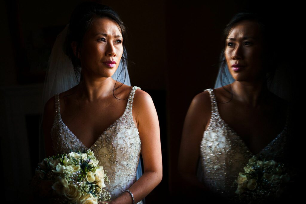 A bride in a beaded wedding gown holds a bouquet of white flowers, gazing thoughtfully into the distance. Her reflection is visible in a nearby mirror, creating a dramatic and introspective moment captured in low lighting. Image by Documentary Wedding Photographer Lewis Bishop Photography.