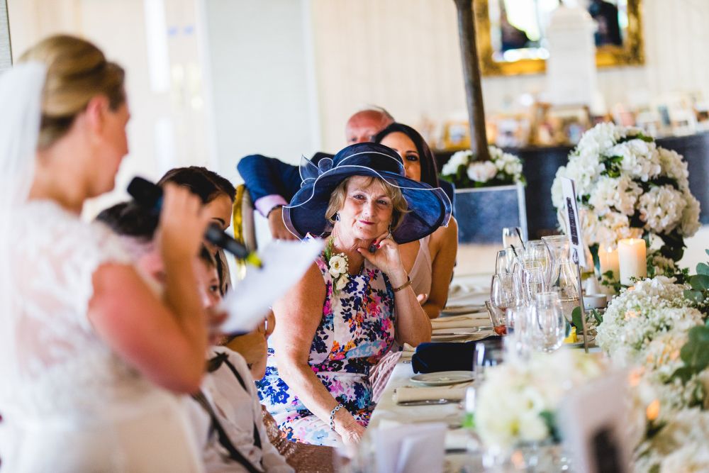 A bride in a white dress speaks into a microphone at a wedding reception. A guest in a floral dress and large blue hat attentively listens, seated at a table decorated with white flowers, candles, and wine glasses. Other guests are seen in the background. Image by Documentary Wedding Photographer Lewis Bishop Photography.